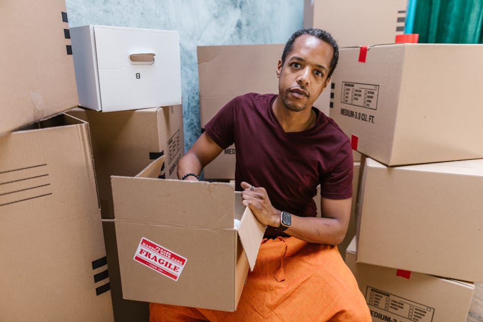 A man wearing a maroon t-shirt and orange pants is kneeling on the floor inside a room filled with packed cardboard boxes, some labeled with extra packaging materials like plastic wrap. He is holding an open cardboard box, preparing it for a home relocation or furniture transport, with a focused expression. The scene shows a typical moving environment with boxes stacked around him, a box marked 'fragile', and a background of a room with light blue walls. The interior lighting is bright and even, highlighting the details of the boxes, packing materials, and the man's clothing. The area appears to be near a doorway or an entry point used for loading items onto a moving van, consistent with professional removals services like those offered by Hackney Man and Van.