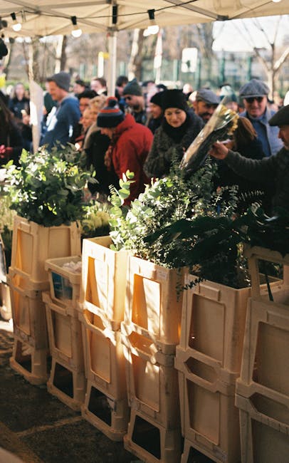 The image shows a busy outdoor market scene during daylight, with a group of people browsing and shopping for plants and flowers displayed on wooden crates and tables under a beige canopy. Several individuals are holding and examining plant arrangements, wrapped in clear plastic, while others look on, creating an active and engaged atmosphere. The market appears to be set up on a paved area, with trees and additional market stalls visible in the background. The bright natural lighting highlights the vibrant greenery and the diverse crowd, with some people dressed warmly, indicating cooler weather. In the context of house removals and moving services offered by Hackney Man and Van, this scene reflects the importance of careful handling and organization during packing and transportation of fragile items like plants, as part of a thorough home relocation process involving furniture transport and on-site loading procedures.
