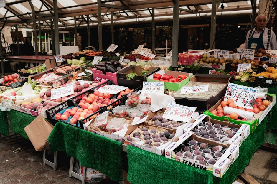 A market stall on a covered outdoor pavement displaying fresh fruits such as peaches, nectarines, and plums arranged in plastic trays and cardboard boxes. The produce is set on a green tablecloth, with handwritten price tags and labels visible on the boxes. In the background, an individual wearing a striped apron and a lanyard stands near the arrangement, with additional boxes of fruit and signs indicating different types of produce. The market setup includes a metal framework supporting the roof, providing shade for shoppers. The scene captures the typical environment of a busy weekly market in Hackney, with vibrant displays of fresh, seasonal fruits prepared for sale, which could be relevant to house removals or moving services when packing or transporting goods in the process of home relocation, as undertaken by Hackney Man and Van.