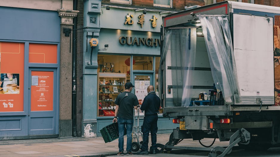 A white moving van with its rear doors open is parked on a city street in front of a commercial property, part of a home relocation or furniture transport process. Inside the van, visible loading equi