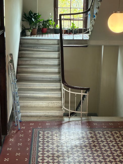 The image shows a staircase inside a residential property, with wooden flooring on the stairs and Victorian-style patterned tiles on the landing at the top. The staircase has a black handrail and white spindles, with a curved section leading to the upper floor. A silver, collapsible step ladder is positioned at the bottom of the stairs, indicating a recent or ongoing move or packing activity. On the landing, there are several potted plants placed near a window, which allows natural light to fill the area. The surrounding walls are painted in neutral tones, and there is a spherical ceiling light fixture on the right side, providing additional illumination. The scene appears to be part of a home relocation process, with a focus on navigating tight staircases and access constraints, consistent with the services offered by Hackney Man and Van and relevant to house removals in the Hackney area.
