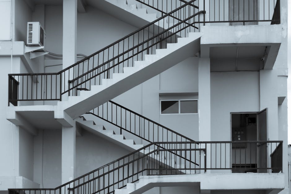 A multi-storey building interior featuring a white concrete staircase with black metal railings on each side, winding through the interior with several landings visible. The space is well-lit with natural light coming from a small rectangular window on the wall, situated between the staircase landings. The staircase appears to be indoors, adjacent to a doorway leading into an apartment or storage area, with smooth white walls and an exposed air conditioning unit mounted on the upper left side. The scene shows the staircase in a static position, with no people or furniture, emphasizing the structural design and spatial arrangement typical of urban residential properties. Hackney Man and Van’s removals services could assist with navigating tight staircases like this during home relocation or furniture transport, especially when handling large items or multiple boxes with packaging materials such as cardboard, plastic wrap, or blankets for protection in confined spaces.