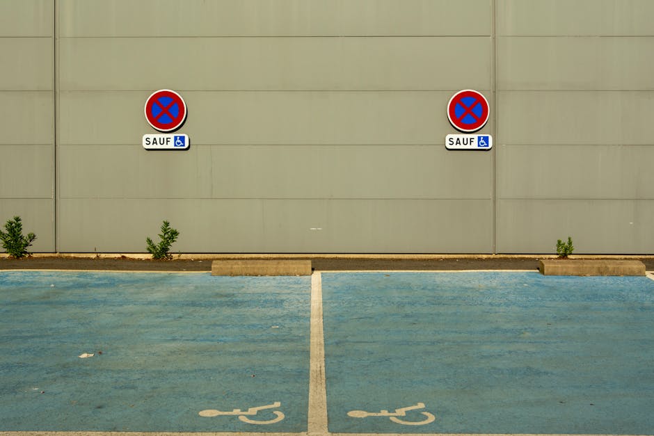 A photograph of a designated parking area with two blue disabled parking bays, each marked with a white wheelchair symbol on the blue surface. A concrete curb separates the parking bays from a plain, 