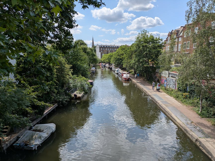 A canal lined with lush green trees on both sides, with several boats moored along the edge, some covered with protective tarps. On the right side, a paved walkway runs parallel to the water, where a few pedestrians are visible, possibly during a home relocation or local move. Parked cars are alongside the path, and behind them, multistory residential buildings with brick facades and large windows are partially obscured by the trees. In the distance, a church spire rises above the cityscape under a partly cloudy sky. The scene captures a calm, urban environment suitable for house removals or furniture transport involving residential streets and waterways, in a setting that reflects typical street-level moving logistics supported by companies such as Hackney Man and Van.