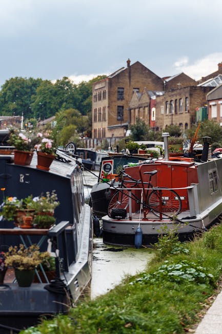 A narrow canal with several residential narrowboats moored along the side, including one with potted plants and decorative items on its roof, and others painted in dark and bright colours. A red bicycle is secured to the roof of a grey boat, which is equipped with a chimney and small windows, indicative of its use as a houseboat. The canal is bordered by a grassy area with some wildflowers and a paved walkway, with a mix of brick and stucco buildings with multiple windows and pitched roofs visible in the background. The sky is partly cloudy with patches of blue, and the scene suggests a calm, residential environment suitable for house removals or moving services, typical of a canal-side relocation process. This image demonstrates the sort of environment that [COMPANY_NAME] might operate in for house removals or furniture transport in the Hackney Wick E9 area, emphasizing the importance of careful packing and transportation during a home relocation or canal-side move.