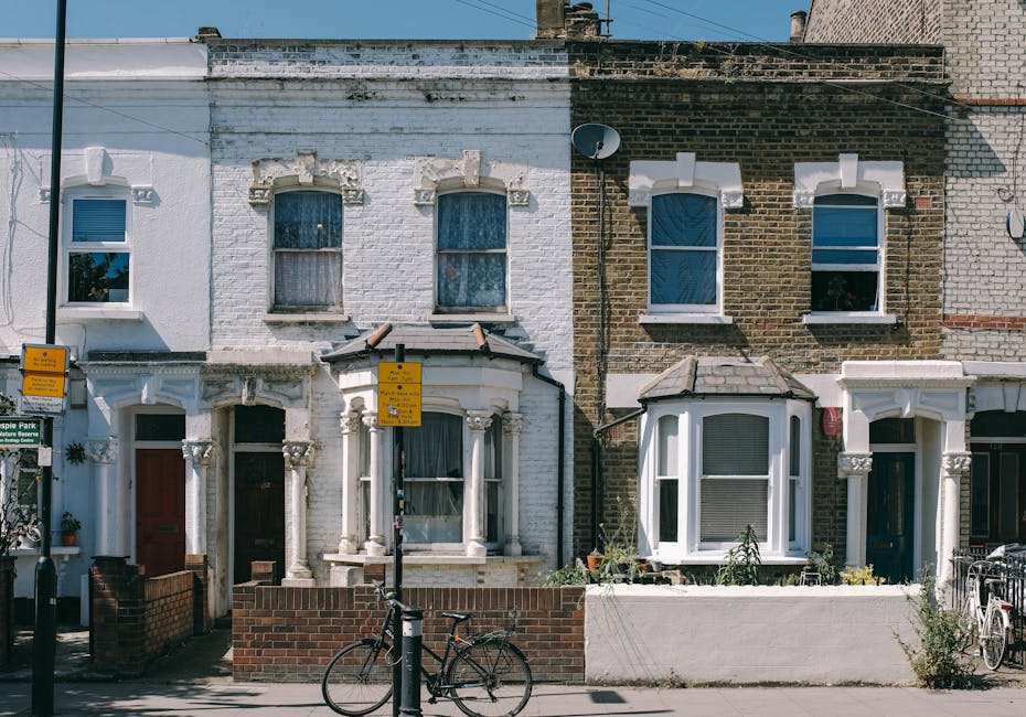 A row of three attached Victorian terraced houses with brick and white-painted facades situated in a residential area. The house on the left features a white brick exterior with decorative stone architraves around a bay window on the ground floor and three sash windows above, all with lace curtains. The middle house has a darker brick facade with white trim around the upper sash windows, and a small bay window with a tiled roof on the ground floor. The doorways are recessed, with the left having a wooden door and the middle a darker door, both with small front yards enclosed by low brick walls. In front of the houses, there is a public sidewalk with a black bicycle leaning against a lamppost, and a yellow street sign with black text mounted on the lamppost. The scene is well-lit with natural daylight, capturing the typical appearance of Victorian-era houses in Stoke Newington N16, suitable for house removals and moving services by Hackney Man and Van.