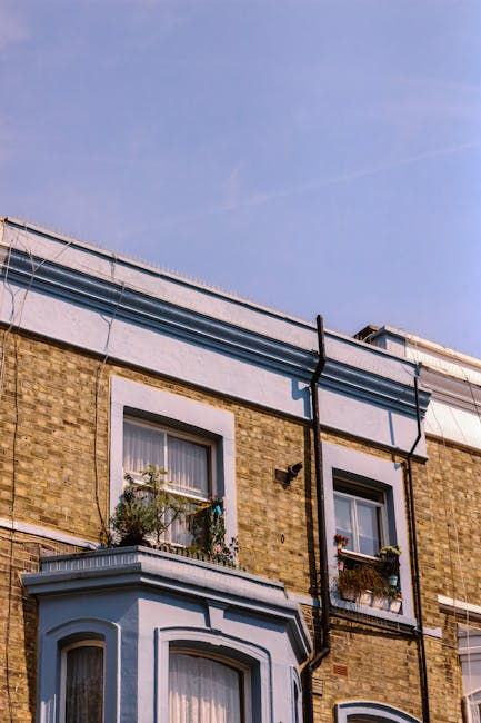 Photograph of a typical Victorian terraced house in Stoke Newington N16, showing a brick facade with two vertically aligned white-framed sash windows, each decorated with flower boxes containing greenery and small plants. The building has a flat roof with a white fascia board along the edge, and black rainwater pipes running down the exterior wall, connecting to gutters at the roofline. The sky above is clear with a blue hue, and the scene is illuminated by natural daylight. This image represents a property suitable for house removals or relocation services, illustrating the type of residential exterior that Hackney Man and Van may service for home moves, including loading furniture and moving boxes through the front door or windows, as part of a comprehensive packing and moving process related to house removals in the area.