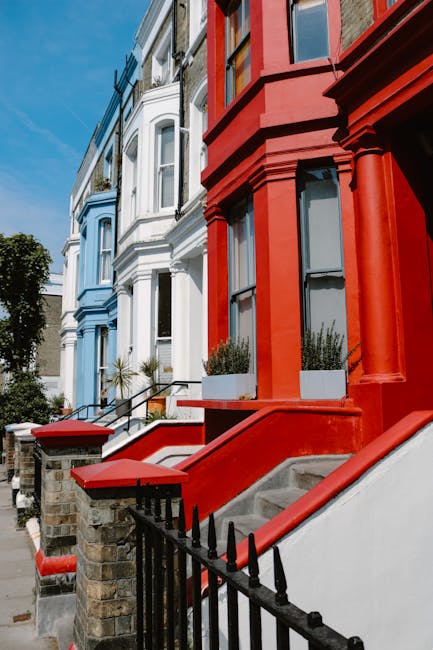 A row of three attached Victorian terraced houses with brick and white-painted facades situated in a residential area. The house on the left features a white brick exterior with decorative stone architraves around a bay window on the ground floor and three sash windows above, all with lace curtains. The middle house has a darker brick facade with white trim around the upper sash windows, and a small bay window with a tiled roof on the ground floor. The doorways are recessed, with the left having a wooden door and the middle a darker door, both with small front yards enclosed by low brick walls. In front of the houses, there is a public sidewalk with a black bicycle leaning against a lamppost, and a yellow street sign with black text mounted on the lamppost. The scene is well-lit with natural daylight, capturing the typical appearance of Victorian-era houses in Stoke Newington N16, suitable for house removals and moving services by Hackney Man and Van.